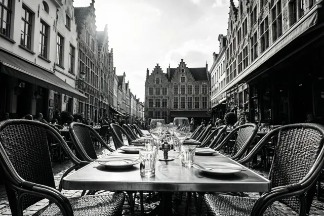 Een zwart-witfoto toont gedekte terrastafels in een smalle Vlaamse straat tussen historische gebouwen onder een bewolkte hemel.