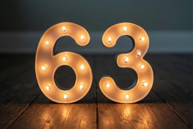Brandende theelichtjes vormen het getal 63 op een donkere houten vloer tegen een donkere achtergrond.