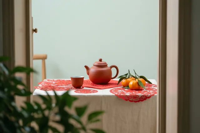 Door een deur- of raamkozijn is een rode aardewerken theepot met een kopje te zien op een tafel met een wit kleed, rode papieren knipsels en mandarijnen met bladeren.