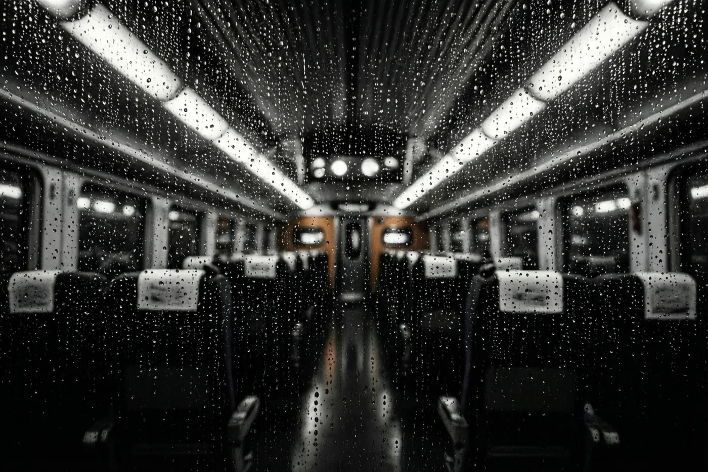 Door een raam met regensporen is een treininterieur te zien met onscherpe plafondlichten en rijen stoelen in een monochroom beeld.