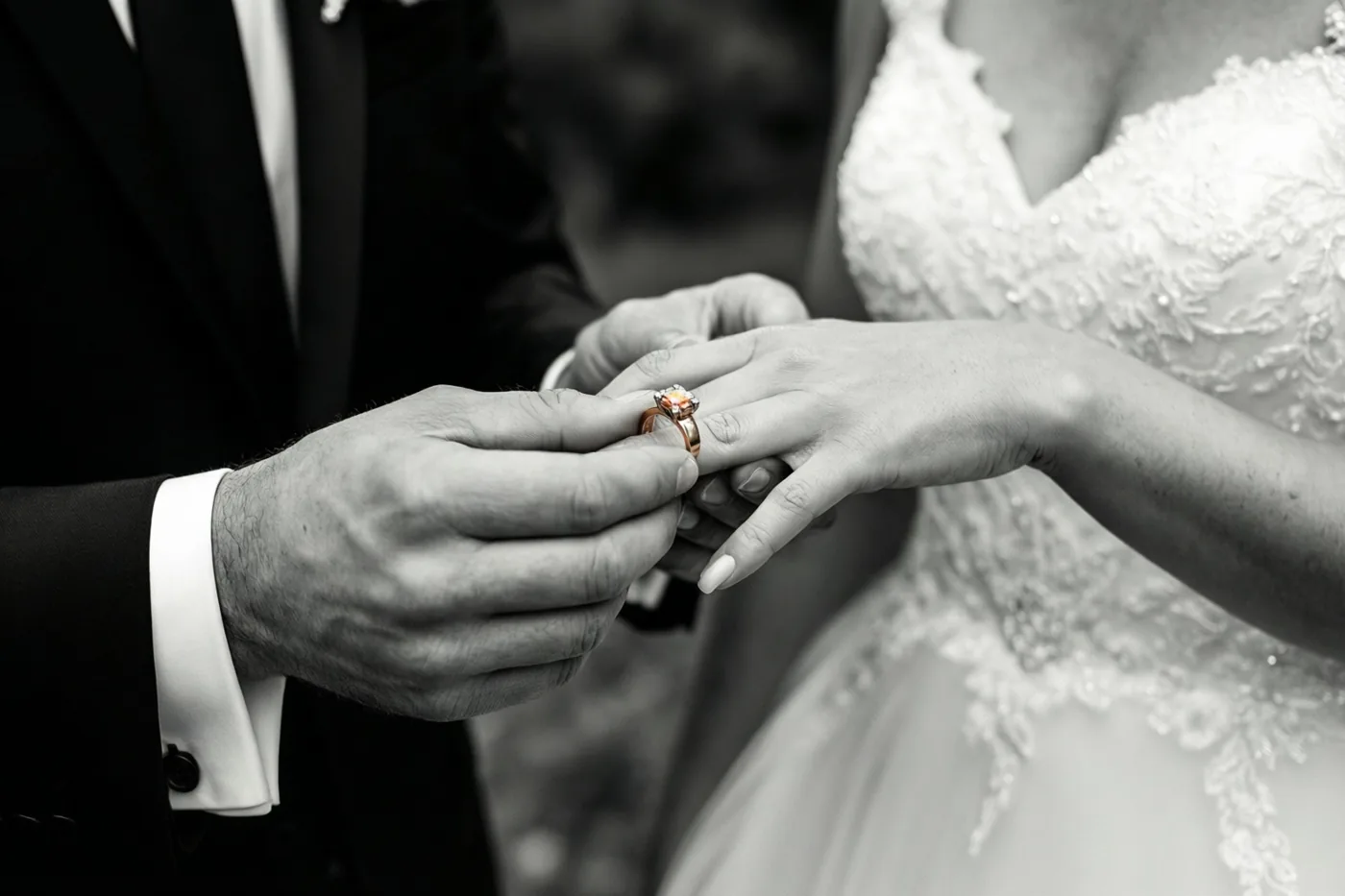Een zwart-witte detailfoto toont de handen van een bruidegom die tijdens een huwelijksceremonie een trouwring aan de vinger van de bruid schuift, met een onscherpe achtergrond.