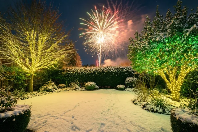 Een besneeuwde tuin is 's nachts verlicht met gele en groene lichtjes in bomen en struiken, terwijl vuurwerk boven de besneeuwde haag uitbarst.