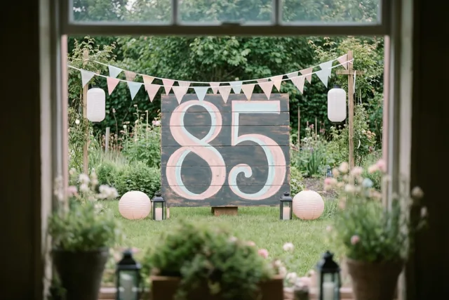 Door een raam is een groene tuin te zien met een groot houten bord met 85, vlaggenlijnen en lantaarns in zachte pasteltinten.