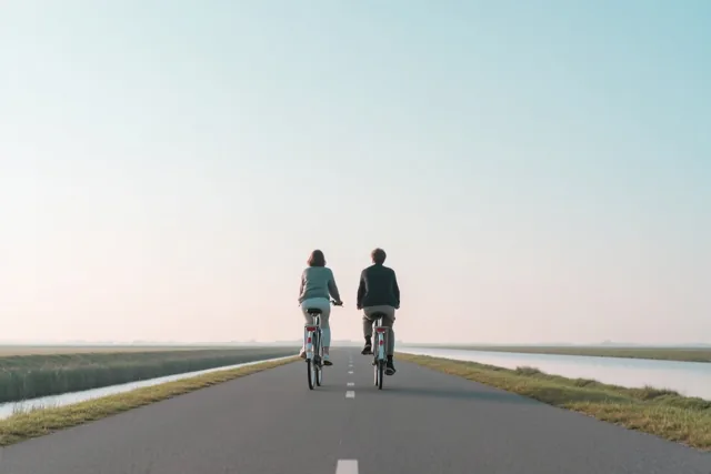 Twee mensen fietsen met hun rug naar de kijker naast elkaar over een rechte asfaltweg door de polders onder een heldere hemel in zacht licht.