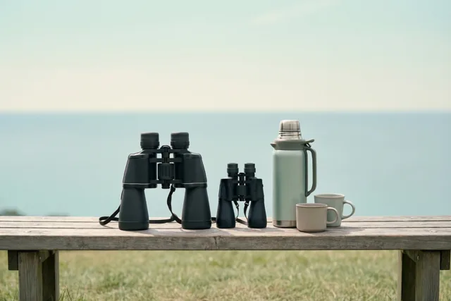 Een grote en een kleine verrekijker liggen op een houten bank naast een thermos en twee mokken, met uitzicht op een kalme zee onder een heldere hemel.