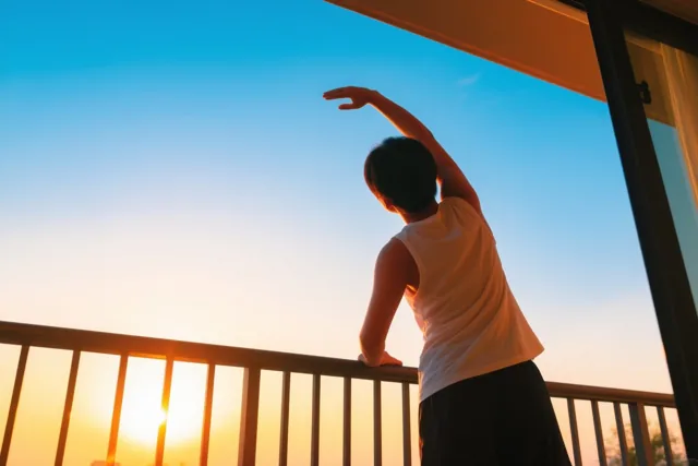 Van achteren zie je een persoon die zich met een geheven arm uitrekt op een balkon, met schaduwen van de reling onder een oranje-blauwe hemel bij zonsopgang.