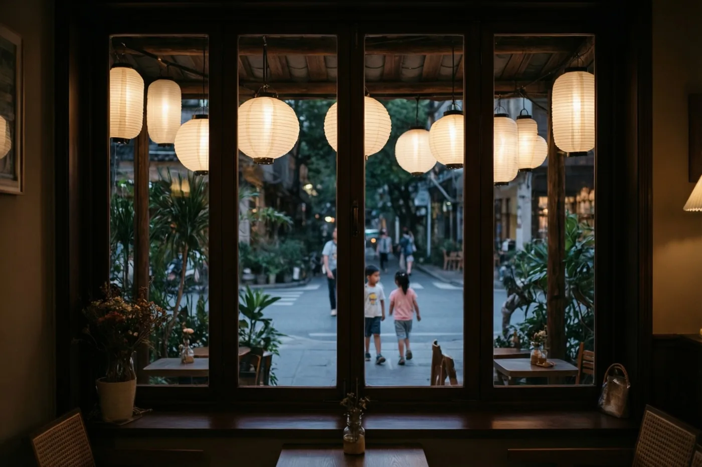 Vanuit een bruin café biedt een groot raam bij valavond zicht op papieren lampionnen buiten en twee kinderen verderop in de straat, in zachtbruine en gele tinten.