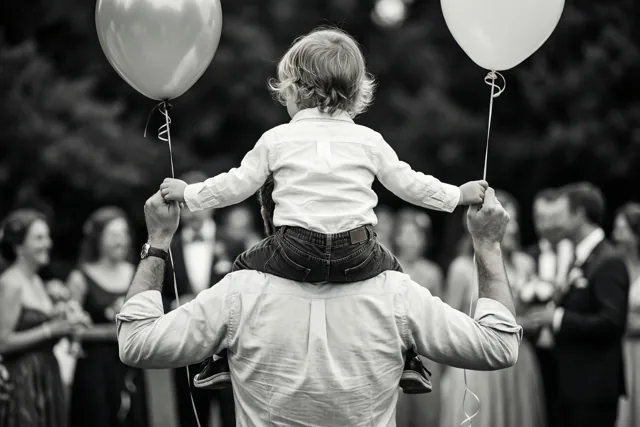 Een zwart-witfoto toont een vader met een kind op zijn schouders tijdens een tuinfeest, terwijl ze allebei ballonnen vasthouden.