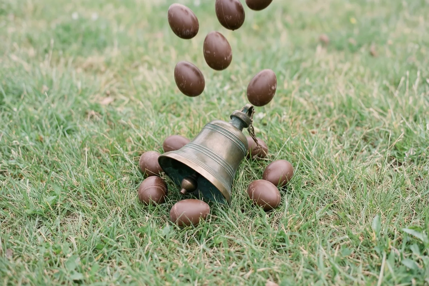Een klein bronzen klokje ligt in groen gras, omringd door chocolade-eieren die uit de lucht lijken te vallen.