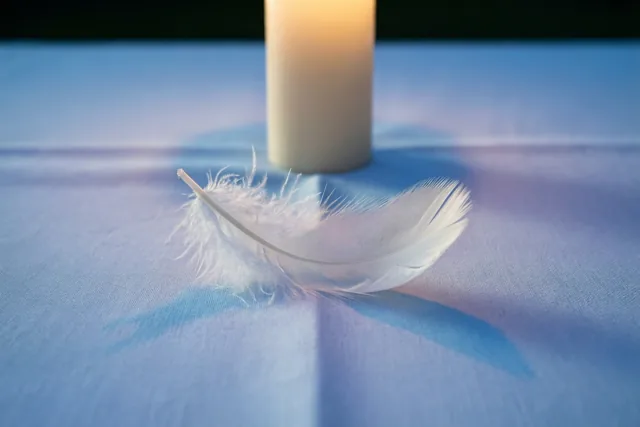 Een witte veer ligt op lichtblauw linnen met een witte doopkaars erachter in zacht licht.