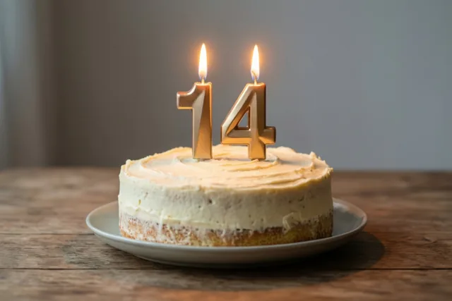 Een zelfgemaakte roomtaart met twee brandende gouden cijferkaarsjes die 14 vormen staat op een wit bord op een houten tafel.