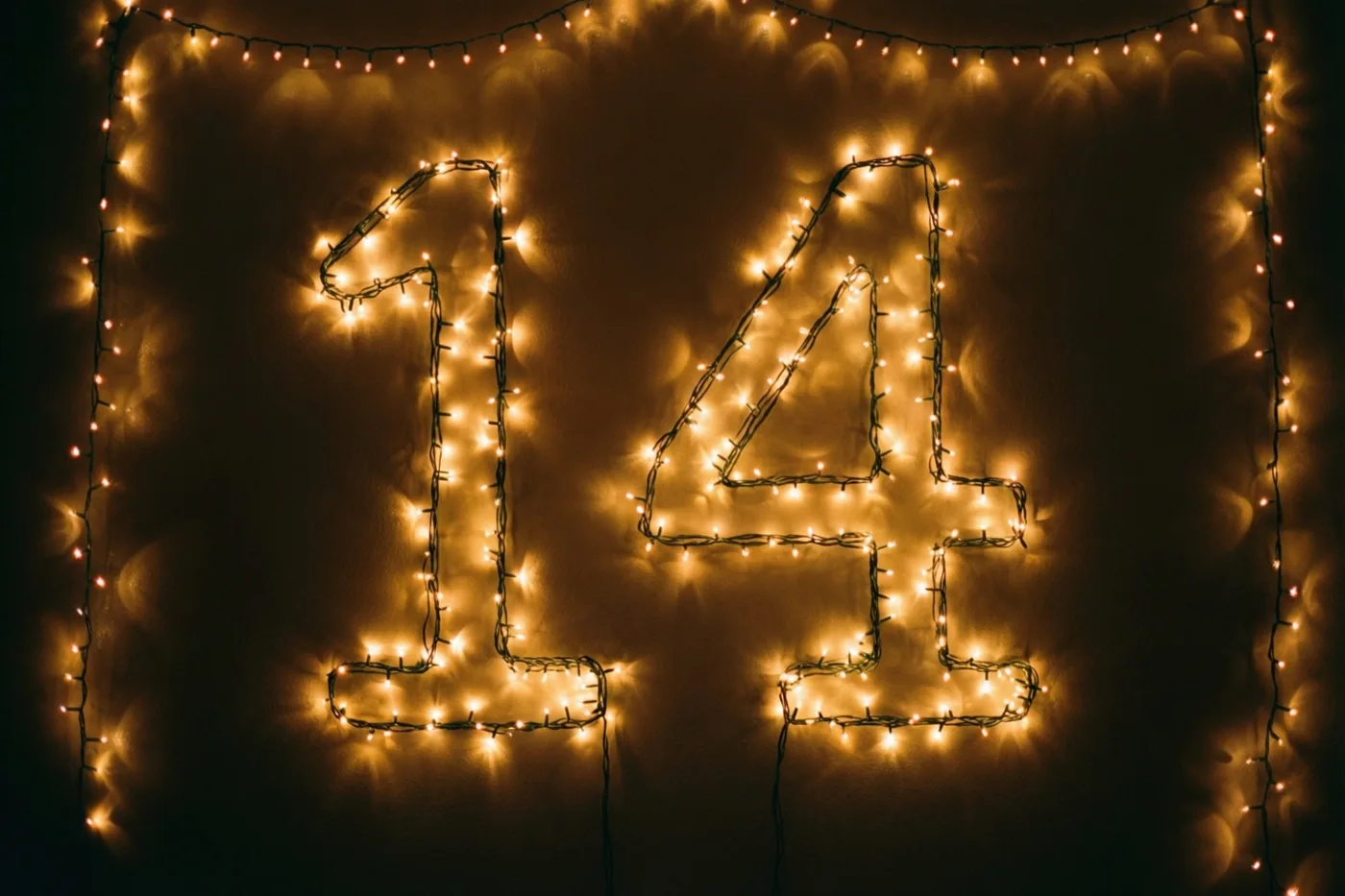 Warme lichtjes vormen 14 tegen een donkere muur, omringd door lichtsnoeren en onscherpe lichtvlekken.
