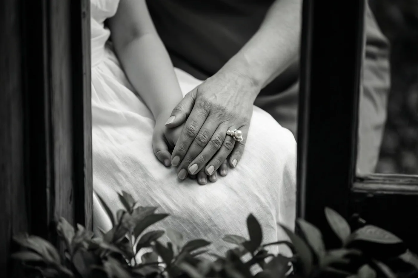 Een zwart-witdetailfoto toont een volwassen hand met een parelring die een kinderhand vasthoudt op wit linnen, omlijst door donkere houten spijlen en onscherp gebladerte op de voorgrond.