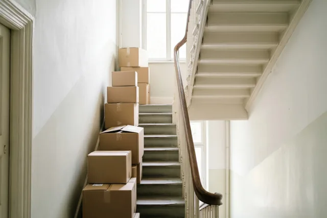 Op elke trede van een smalle trap in een oud herenhuis staan verhuisdozen, met bovenaan een raam dat daglicht binnenlaat.