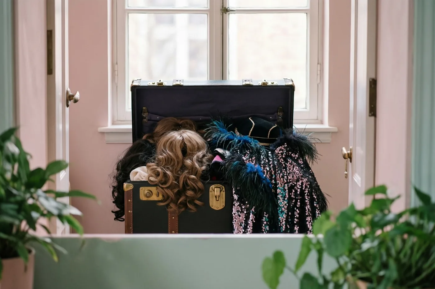 Een open zwarte koffer vol kleurrijke pruiken, veren en capes met pailletten staat voor een raam in een kamer met roze muren en groene planten op de voorgrond.