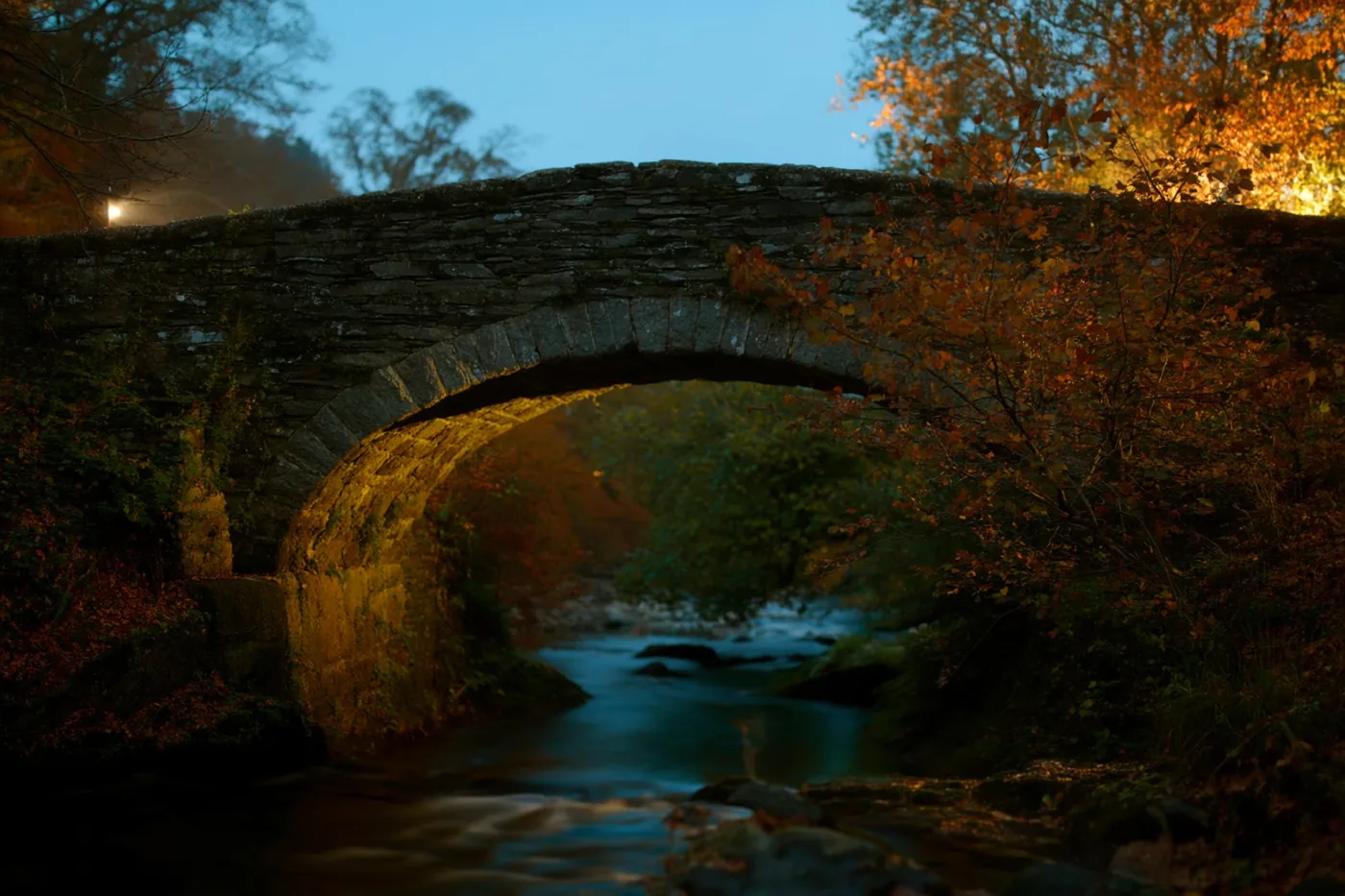 Een stenen boogbrug over een beek en bomen met oranje en rode bladeren worden bij schemering warm verlicht onder een blauwe lucht.