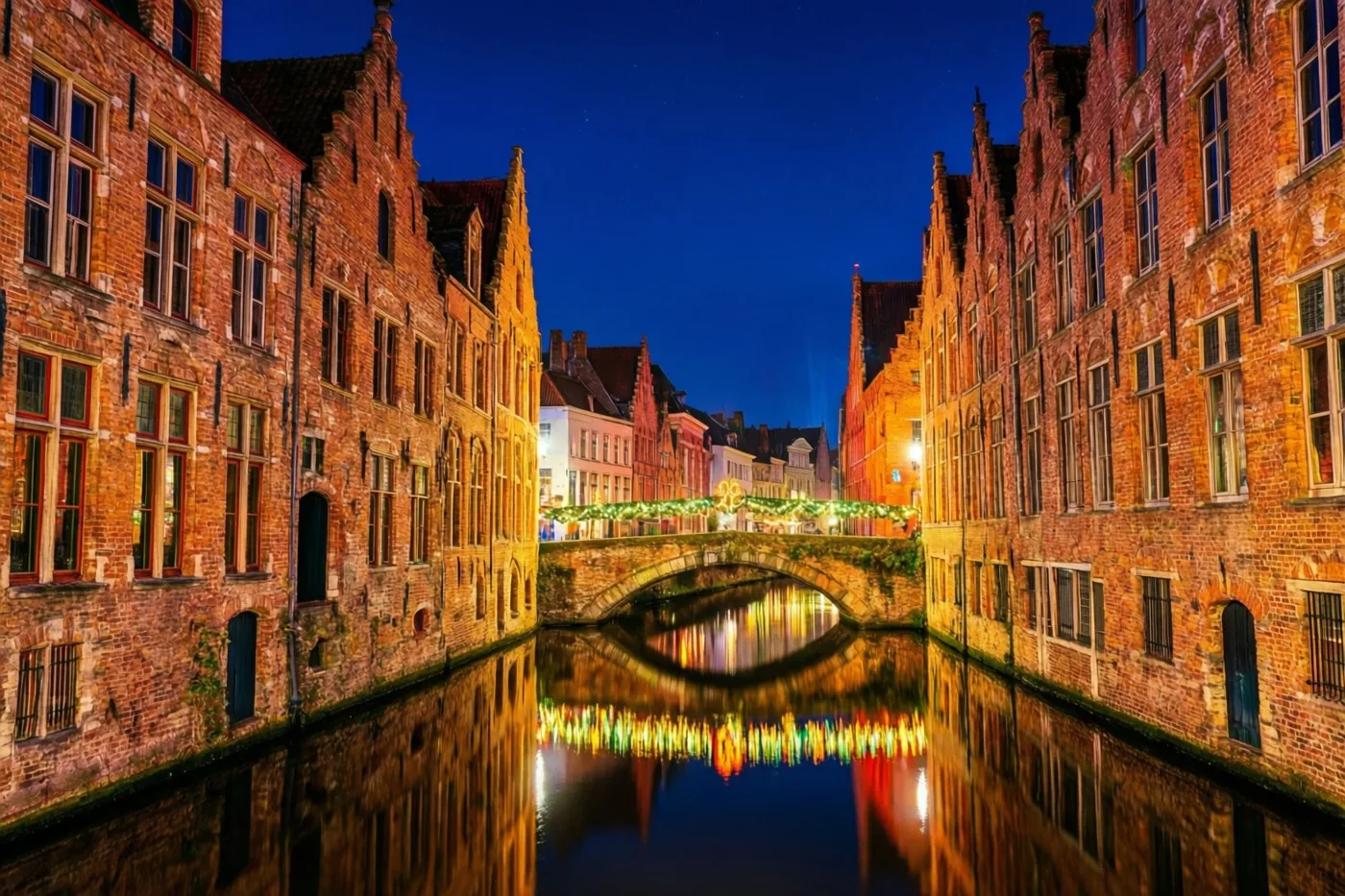 Een smalle waterloop in een oud Vlaams stadje weerspiegelt 's nachts historische bakstenen gevels en warme lichtjes aan een stenen brug.
