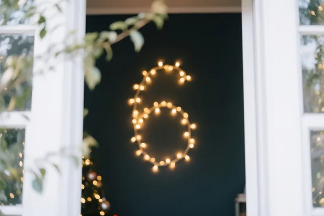 Door een raam met een wit kozijn is een cijfer 6 van warme lichtjes te zien op een donkere muur, met onscherp groen op de voorgrond.
