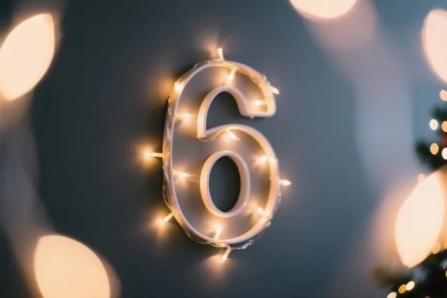 Een cijfer 6 van warme lichtjes hangt aan een donkere muur, omringd door onscherpe feestlichtjes in gele en blauwe tinten.