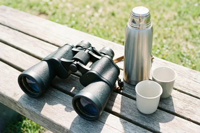 Een zwarte verrekijker ligt op een verweerde houten bank naast een zilverkleurige thermos en twee witte kopjes, met zonnig groen gras op de achtergrond.