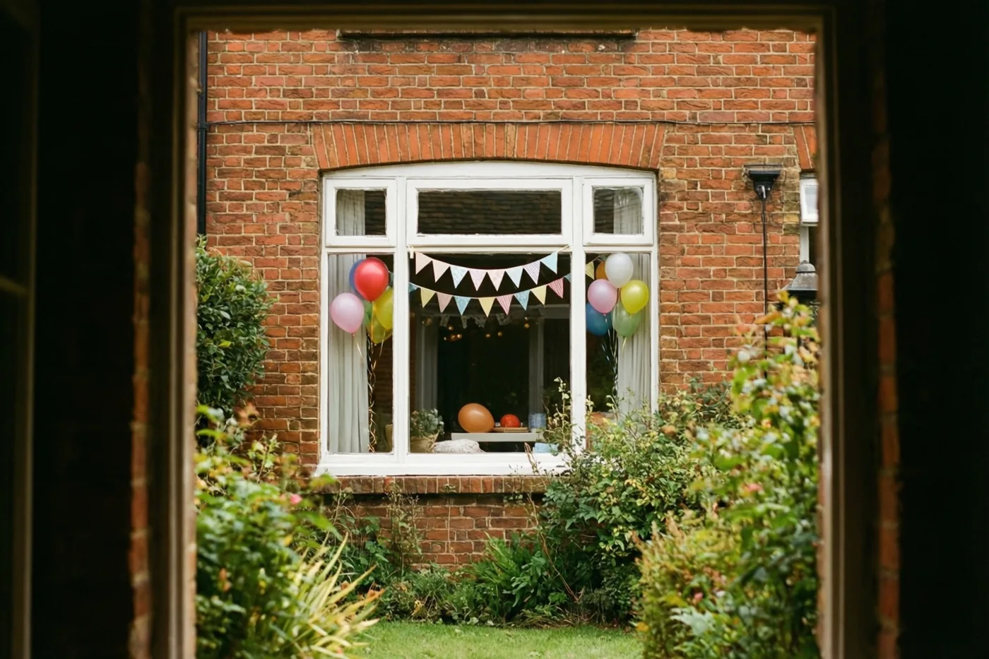Door een houten omlijsting is een raam van een bakstenen huis te zien, versierd met kleurrijke papieren vlaggetjes en ballonnen, met groen in de tuin.