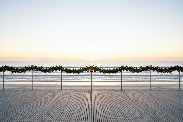 Een houten wandelpromenade aan zee heeft bij schemering een leuning met kerstslingers en versieringen, tegen een achtergrond in zachtblauw, oranje en wit.