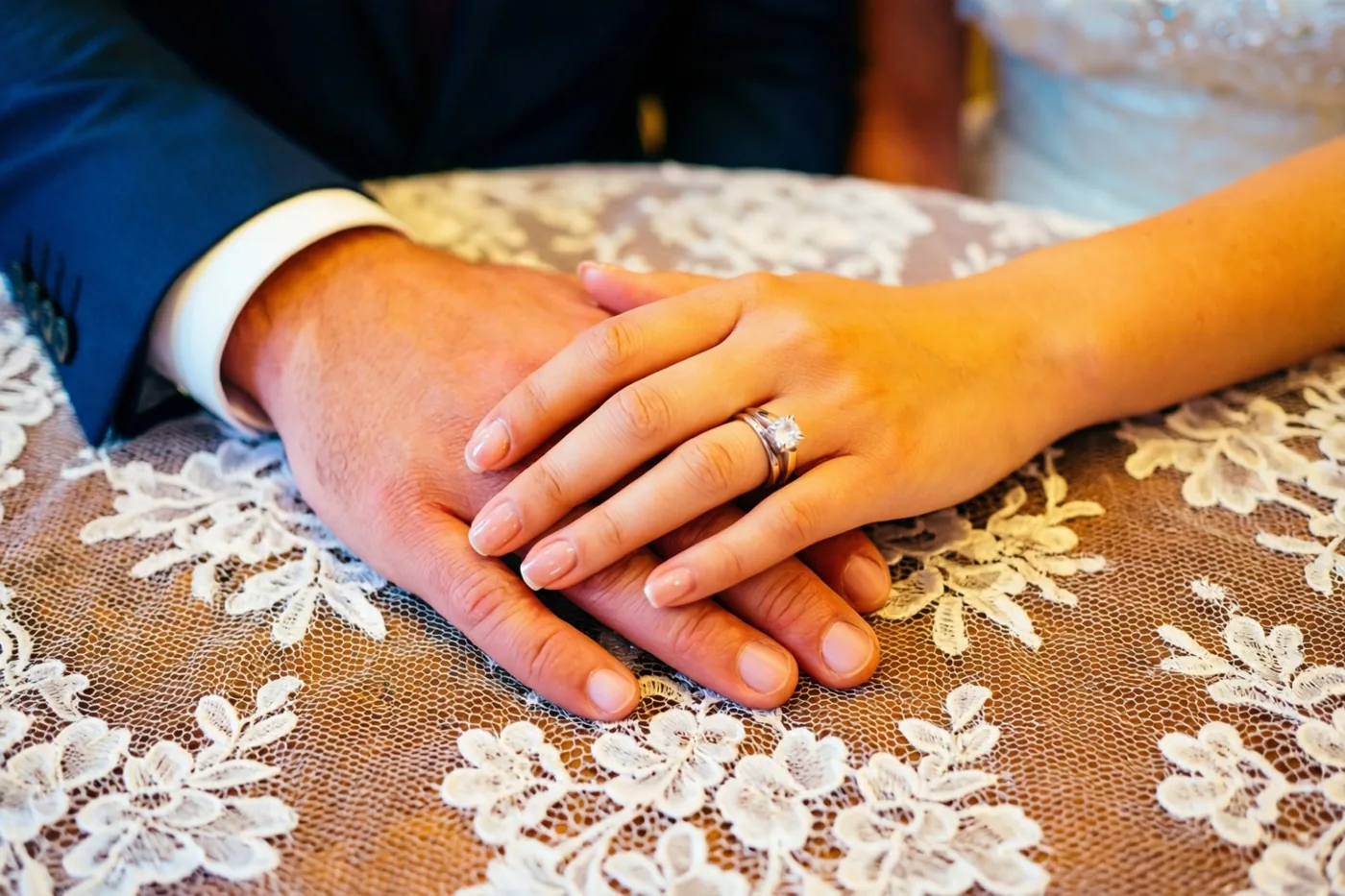 Een mannenhand en een vrouwenhand zijn verstrengeld boven een wit tafelkleed met bloemenkant, met een opvallende diamanten verlovingsring aan de vrouwenhand in zacht warm licht.