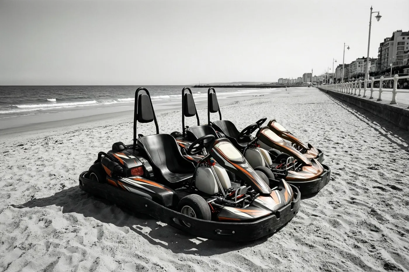 Vier gocarts staan op een rij op een breed zandstrand naast een zeepromenade, in zwart-wit met oranje accenten op de gocarts.