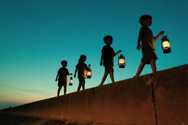 Vier kinderen lopen achter elkaar over een betonnen dijk met elk een brandende lantaarn onder een blauwgroene tot lichtgele schemerhemel.