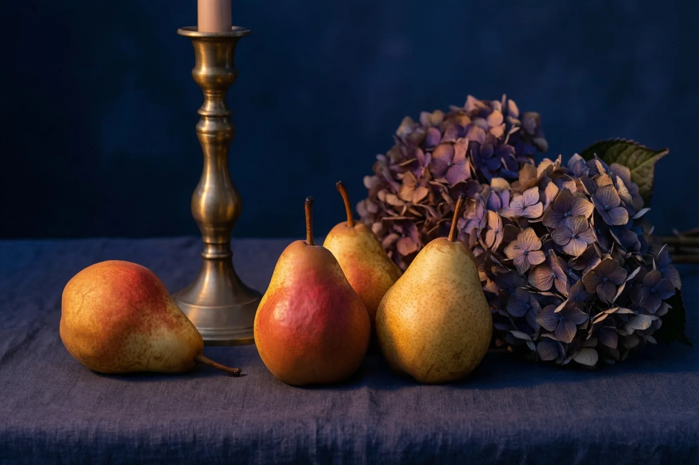 Vier peren, een messing kandelaar met een kaars en een boeket gedroogde hortensia's staan op donkerblauw linnen tegen een diepblauwe achtergrond.