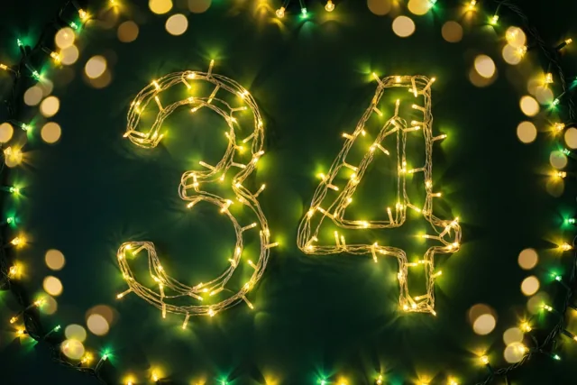 Het getal 34 is gevormd uit warme lichtslingers tegen een donkere muur, met gouden en groene onscherpe lichtjes.