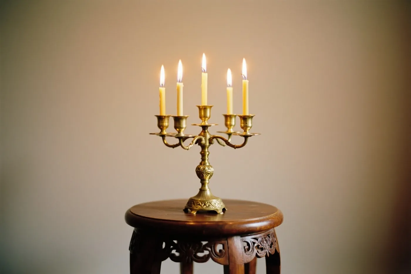 Een messing kandelaar met vijf brandende kaarsen staat op een houten krukje met snijwerk voor een effen muur.