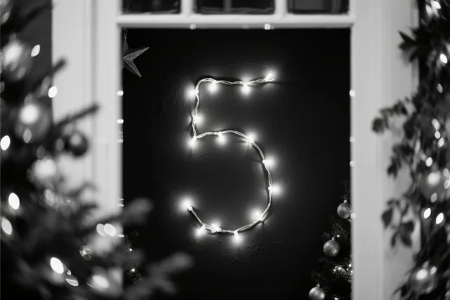 Een cijfer 5 van gloeiende lichtjes hangt aan een donkere muur, omlijst door onscherpe kerstboomtakken en lichtvlekken.