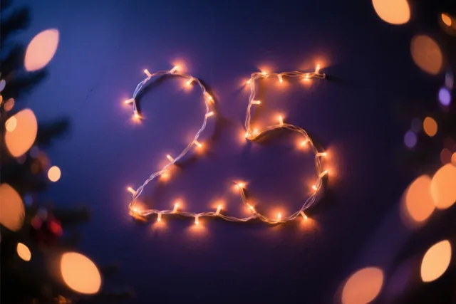 Het getal 25 is gevormd met warme lichtjes op een donkerblauwe muur, met onscherpe feestlichtjes ervoor en erachter.