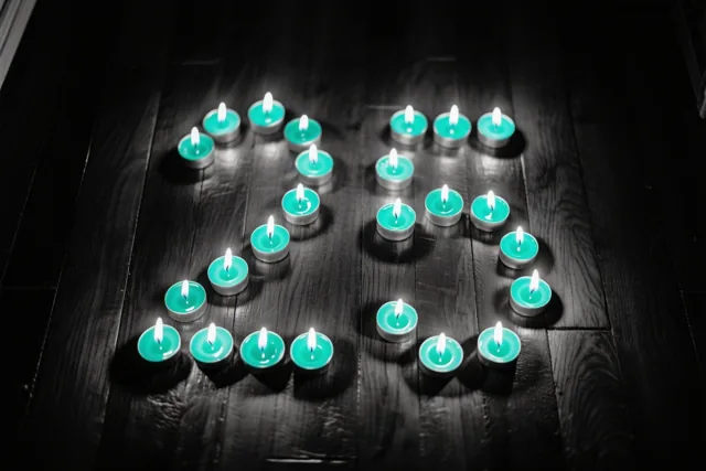 Brandende theelichtjes vormen 25 op een donkere houten vloer, met blauwgroene en zwarte tinten rondom de warme gloed.