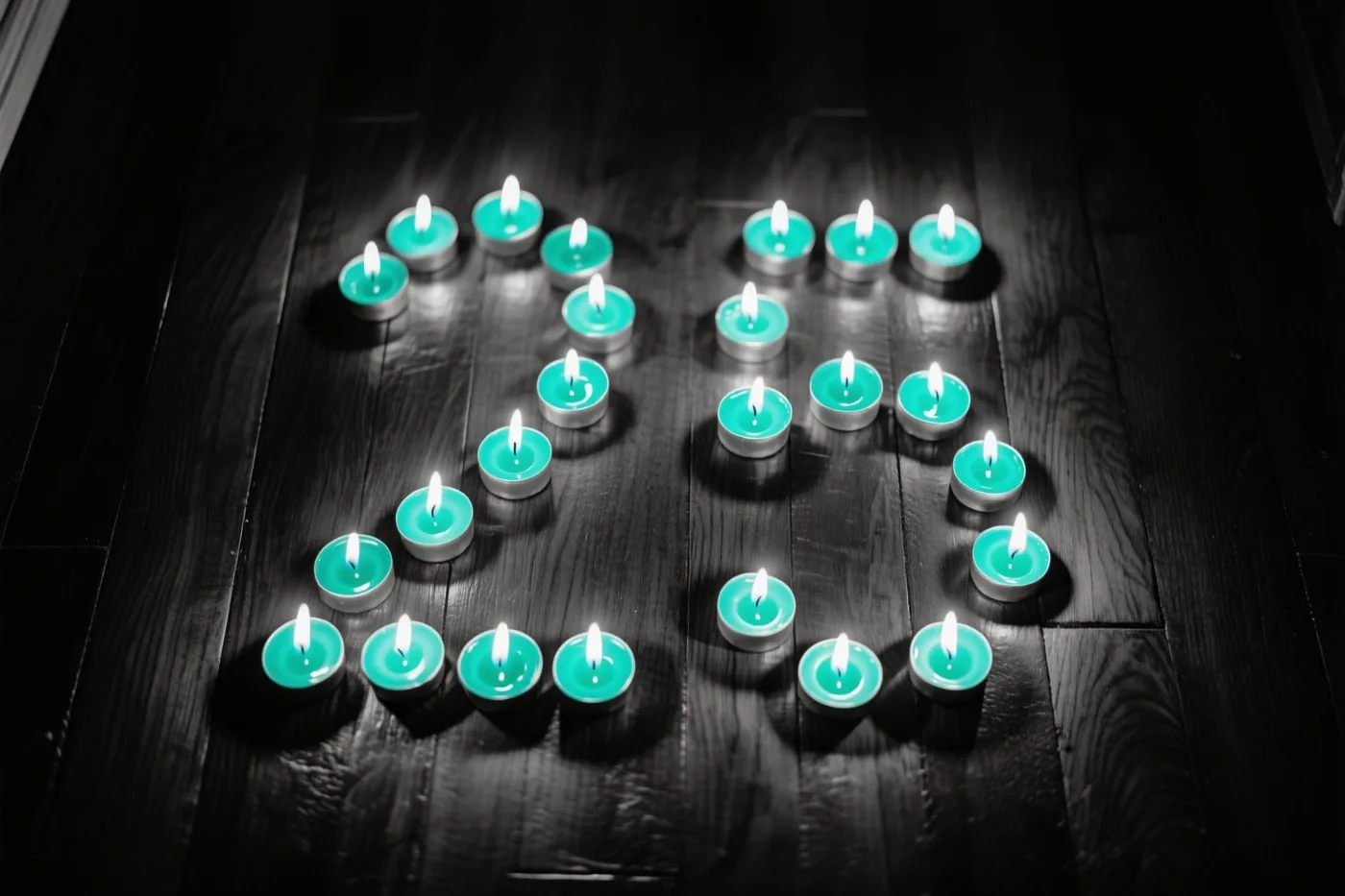 Brandende theelichtjes vormen 25 op een donkere houten vloer, met blauwgroene en zwarte tinten rondom de warme gloed.