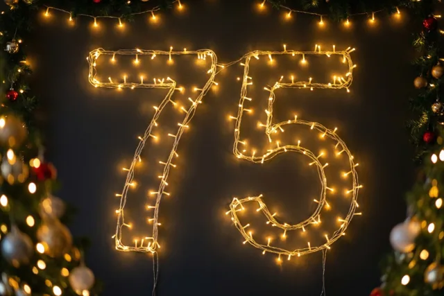 Warme lichtjes vormen 75 tegen een donkere muur, omlijst door wazige kerstboomversiering en lichtjes in gouden tinten.