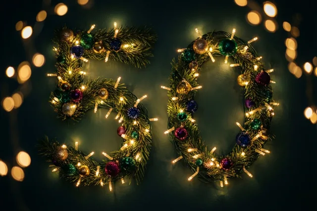Groene dennentakken met kleurrijke versieringen en warme lichtjes vormen 50 tegen een donkere muur met onscherpe lichtjes op de achtergrond.