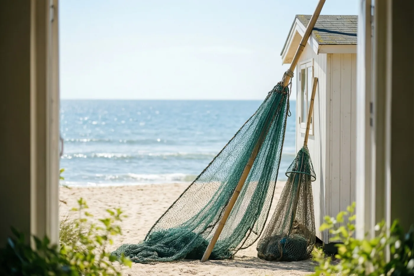 Een groot en een klein visnet leunen tegen een witte strandcabine op een zandstrand, met de zee op de achtergrond in fel zonlicht.