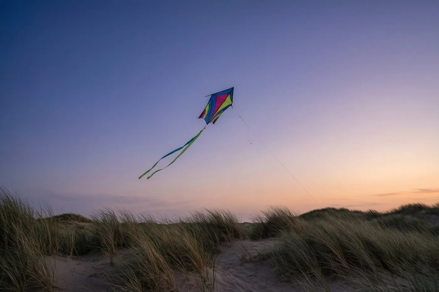 Een kleurrijke vlieger met lange linten vliegt hoog boven zandduinen met hoog gras, onder een lucht die van diepblauw naar zachtoranje aan de horizon verloopt.