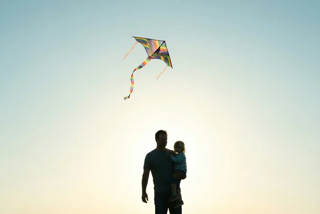 Een vader houdt een kind vast terwijl ze een kleurrijke vlieger oplaten, als silhouetten tegen een lucht die van lichtgeel naar lichtblauw overgaat.