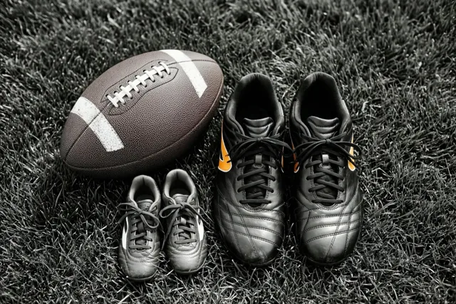 Een voetbal en twee paar voetbalschoenen liggen op het gras in een grotendeels zwart-witbeeld, met gekleurde logo's op de grotere schoenen.