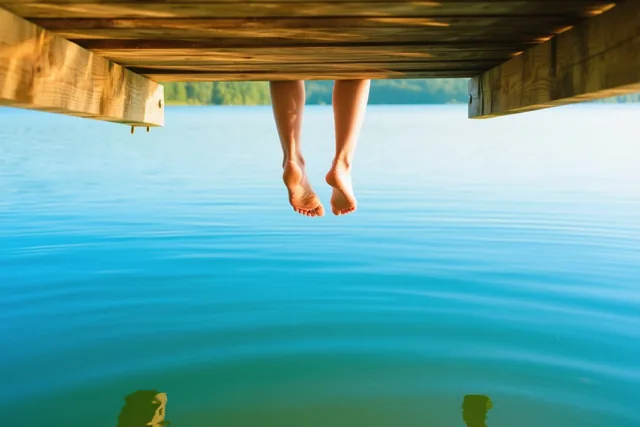Blote voeten hangen onder een houten steiger boven een rustig blauw meer in warm zonlicht, met een groene oever in de verte.