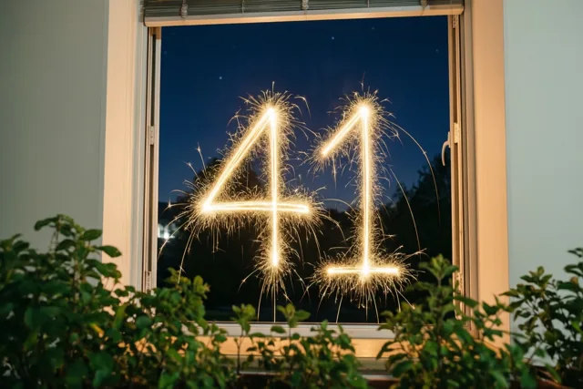 Gouden vonkensporen vormen 41 op een raam bij nacht, omlijst door het raamkozijn en kamerplanten tegen een diepblauwe lucht.