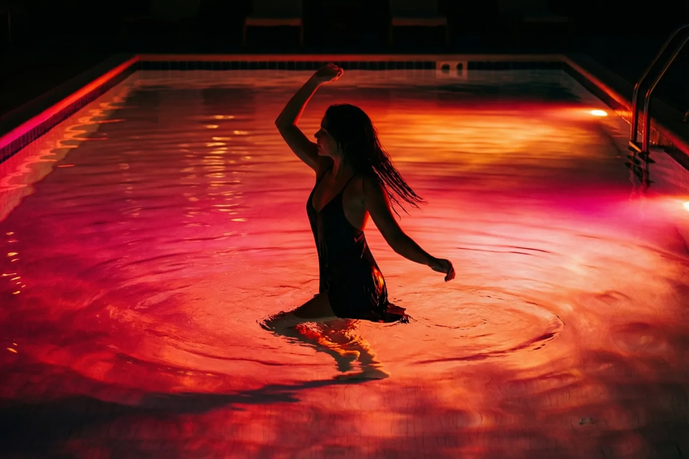 Het silhouet van een vrouw in een zwembad steekt 's nachts af tegen rode en oranje onderwaterverlichting die op het water weerspiegelt.