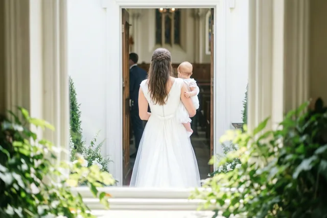 Een vrouw in een witte jurk draagt een baby in een witte doopjurk door een open kerkdeur, met groene bladeren op de voorgrond.