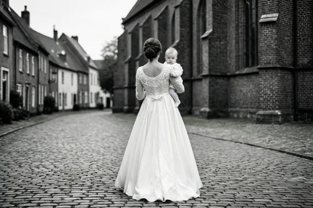 Een vrouw in een lange witte jurk draagt een baby in het wit over een kasseistraat naar een bakstenen kerk, van achteren gefotografeerd in zwart-wit.