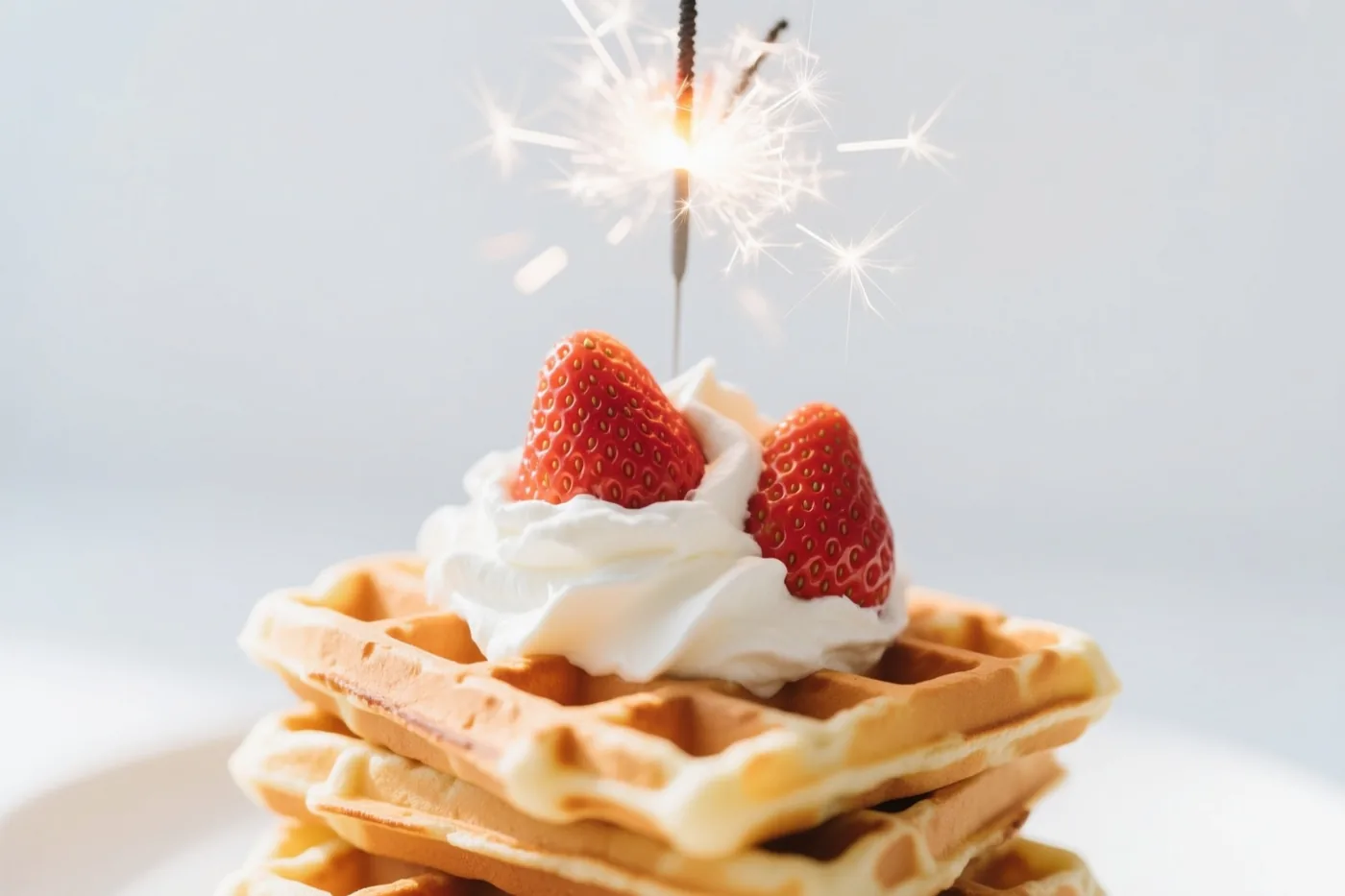 Een stapel goudbruine Belgische wafels met slagroom, twee verse aardbeien en een brandend sterretje staat tegen een effen lichtgrijze achtergrond.