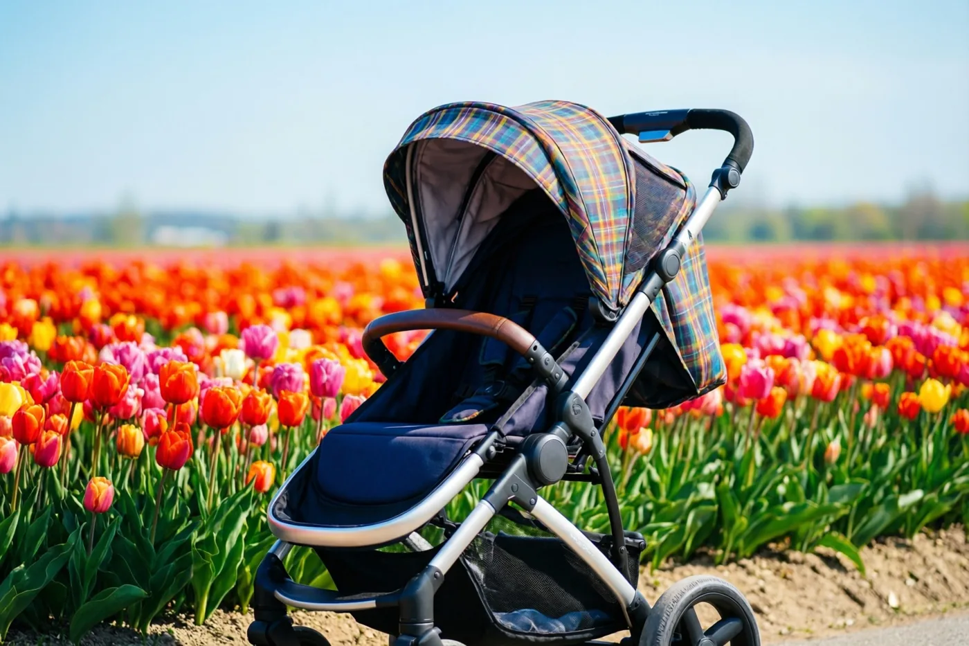 Een wandelwagen met een geruite kap staat naast een veld vol kleurrijke tulpen onder een lichtblauwe lucht.