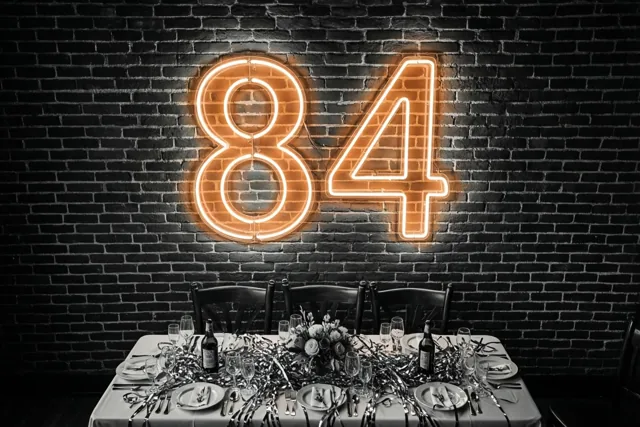 Een warm verlichte neon-84 hangt op een donkere bakstenen muur boven een feestelijk gedekte tafel met slingers en servies.
